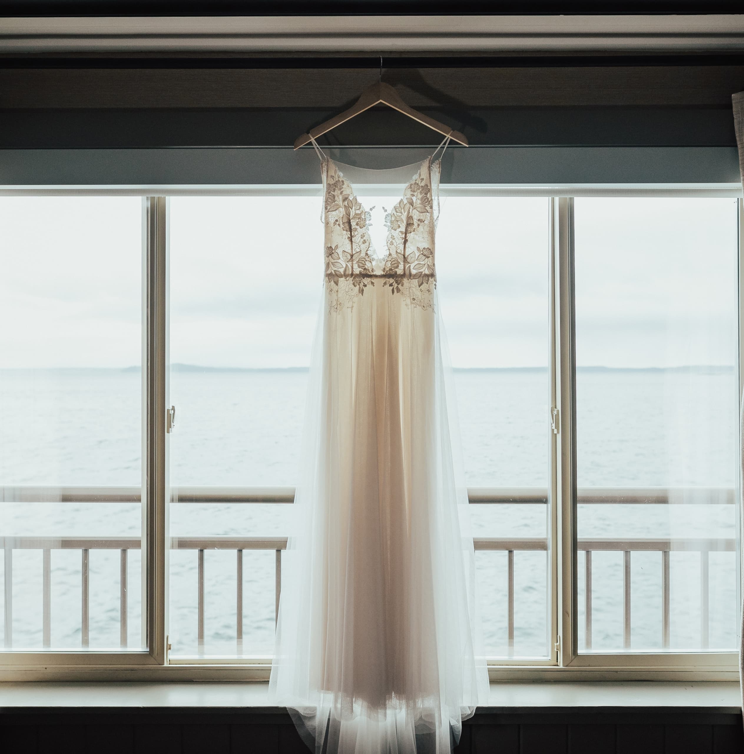 A beautiful white wedding gown hanging in a room with large bay windows overlooking the ocean